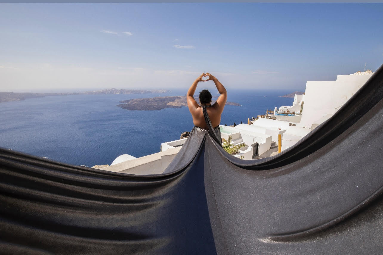 A traveler in a flowing gown making a heart with her hands above the Santorini caldera in Greece