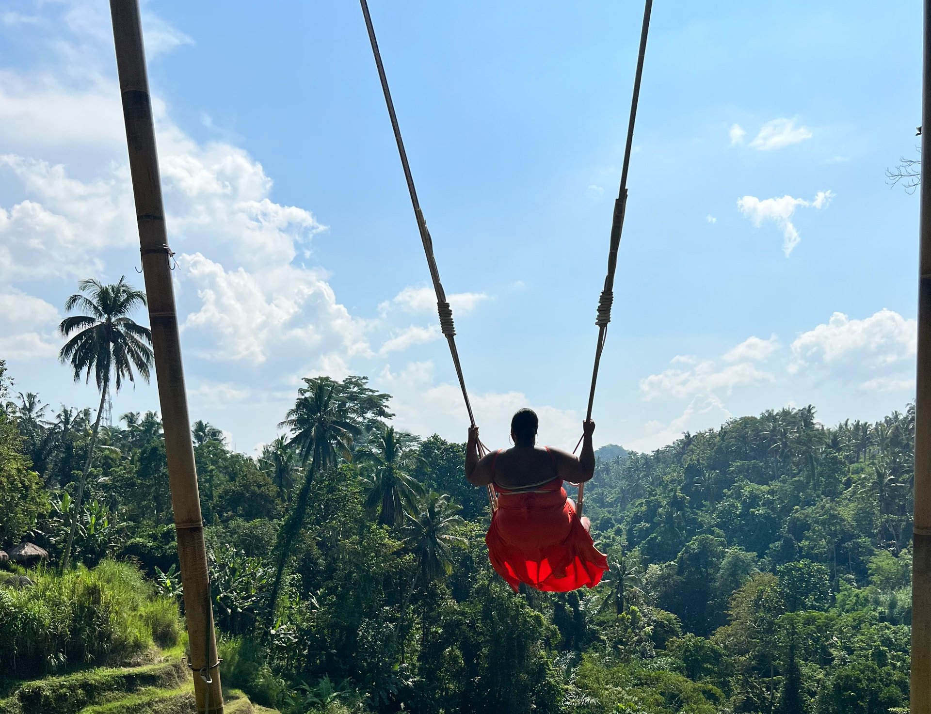 A traveler in a flowing red dress on the Bali jungle swing above palm treetops